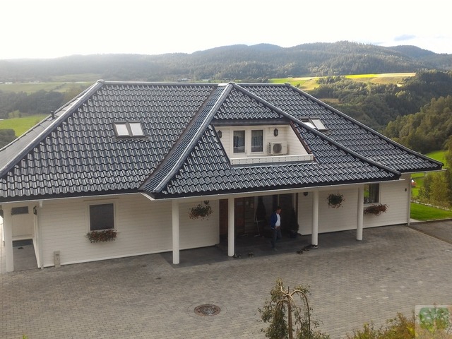 Biały dom z ciemną dachówką na tle zielonych wzgórz. Widoczne okna dachowe i dekoracje kwiatowe. Na podjeździe stoi mężczyzna.