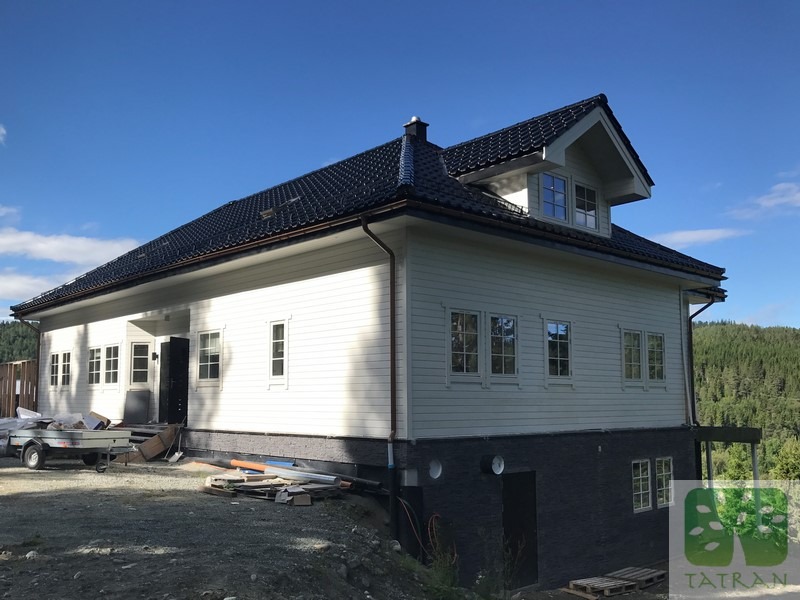 Biały dom z ciemnym dachem i widocznym fragmentem podpiwniczenia, otoczony zielenią. Widoczny fragment przyczepki i materiałów budowlanych.