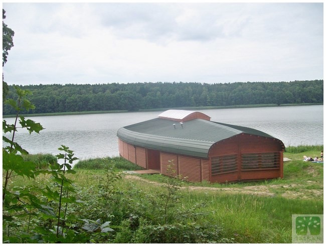 Nietypowy, drewniany budynek z zaokrąglonym dachem, położony nad jeziorem, otoczony zielenią. Widoczne elementy konstrukcyjne i naturalne otoczenie.