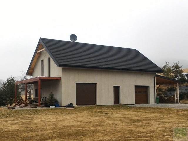 Dwukondygnacyjny budynek z płaskim dachem i dwoma bramami garażowymi, z drewnianą konstrukcją zadaszenia po bokach, na tle pochmurnego nieba i pagórkowatego terenu.