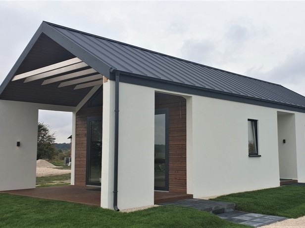 Nowoczesny dom z białymi ścianami i ciemnoszarym dachem z blachy na tle zielonego trawnika. Widoczne drewniane elementy wykończeniowe i minimalistyczna architektura.
