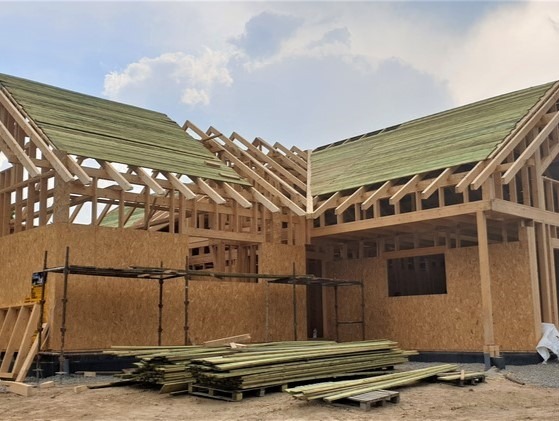 Konstrukcja domu szkieletowego w trakcie budowy: drewniany szkielet, płyty OSB, częściowo pokryty dach, stos drewna na ziemi. Ujęcie z dołu, pochmurne niebo.