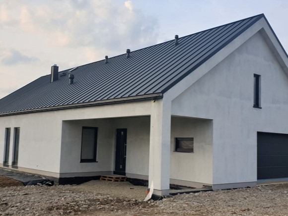 Nowoczesny, parterowy dom z ciemnoszarą blachą na dachu i białymi ścianami. Widoczny garaż, okna i drzwi w ciemnej tonacji. Teren wokół domu w trakcie zagospodarowania.