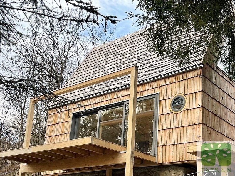 Nowoczesny dom szkieletowy obłożony drewnianym gontem, z tarasem i okrągłym oknem, na tle drzew. Widoczny fragment logo firmy.
