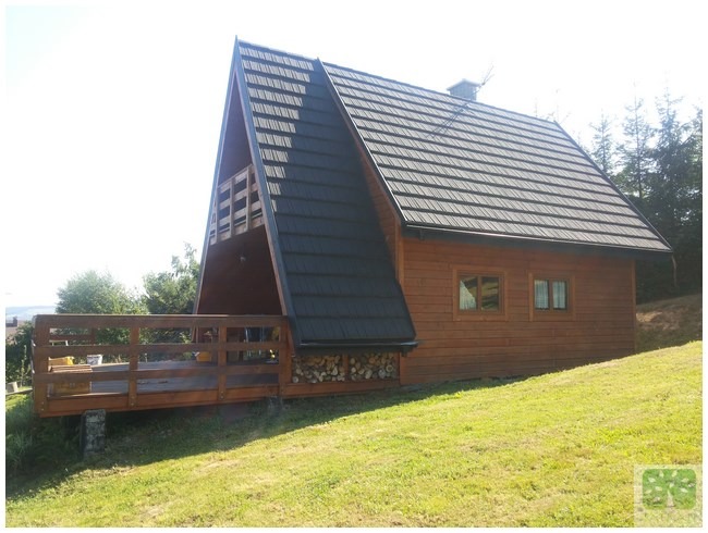 Drewniany dom w stylu A-frame z ciemnym dachem, widoczny z boku, z drewnianym tarasem i stosem drewna pod nim, na tle zielonego wzgórza i drzew.