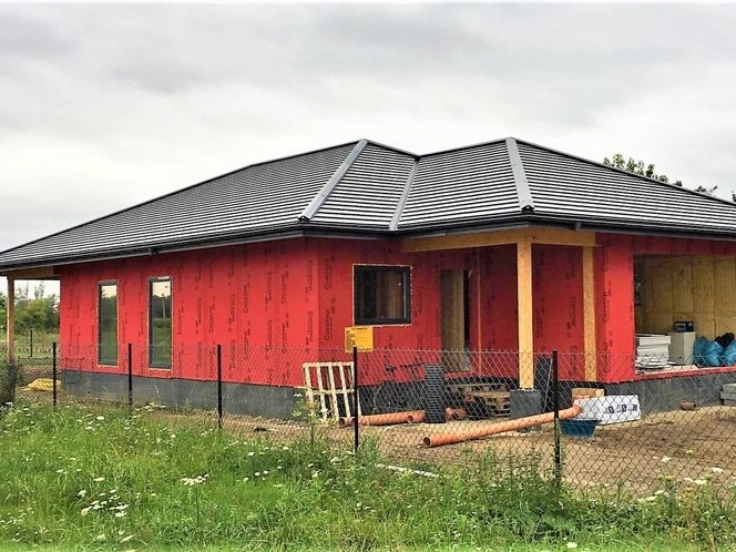 Budowa parterowego domu z czerwonymi panelami ściennymi i ciemnym dachem, widok z zewnątrz przez ogrodzenie z siatki, w tle zielona trawa i pochmurne niebo.