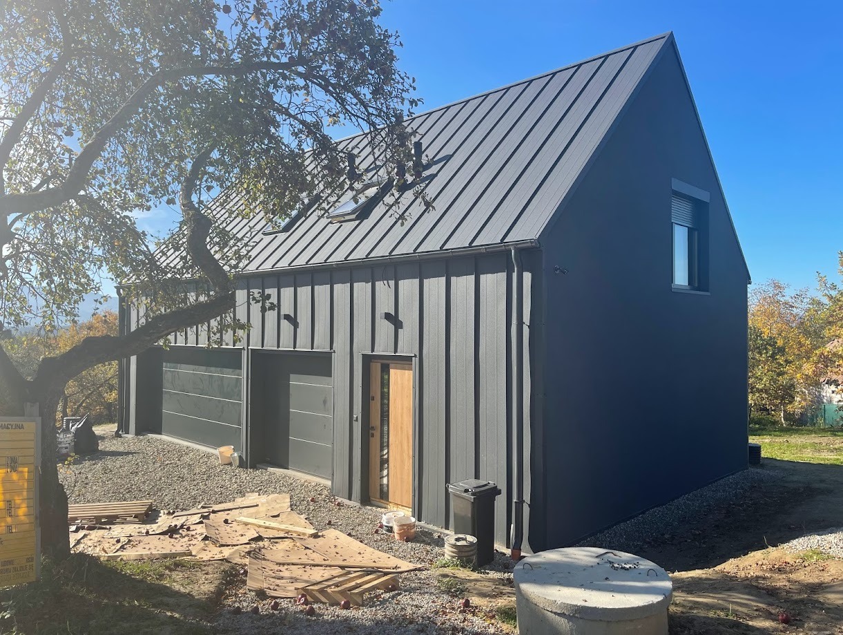 Nowoczesny, grafitowy dom z blachy na rąbek stojący, z dwoma bramami garażowymi i drewnianymi drzwiami wejściowymi, na działce z drzewem i elementami wykończeniowymi.