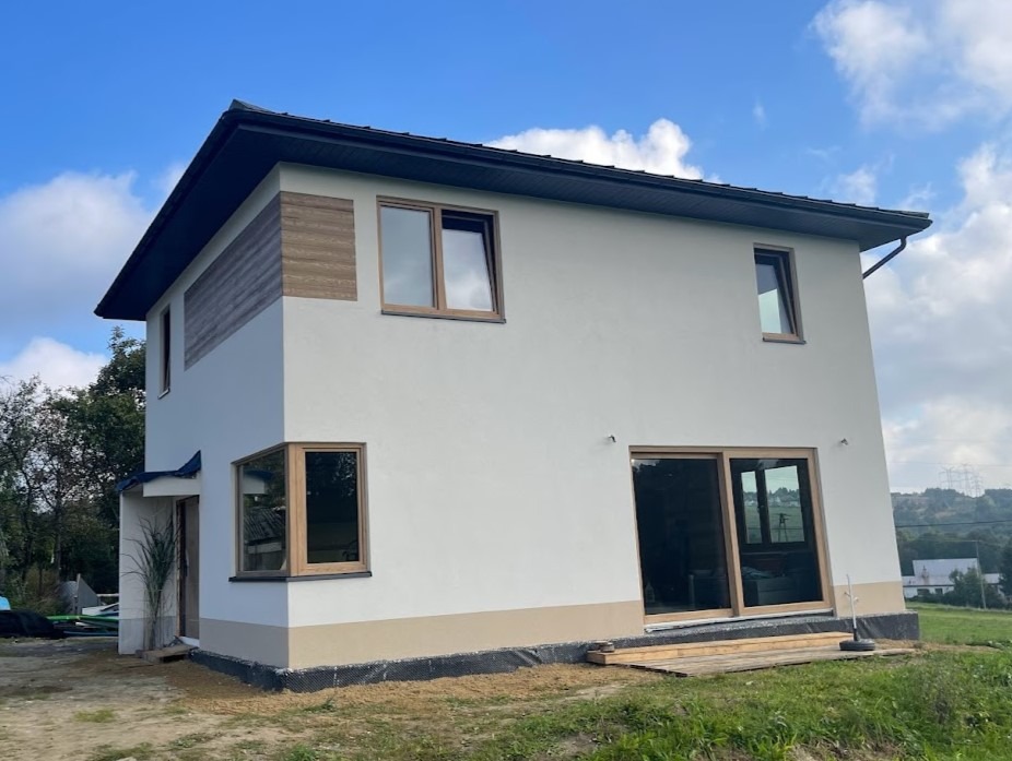 Nowoczesny, dwukondygnacyjny dom z jasną elewacją, drewnianymi akcentami i dużymi oknami, otoczony zielenią. Widok z dołu, podkreślający bryłę budynku.