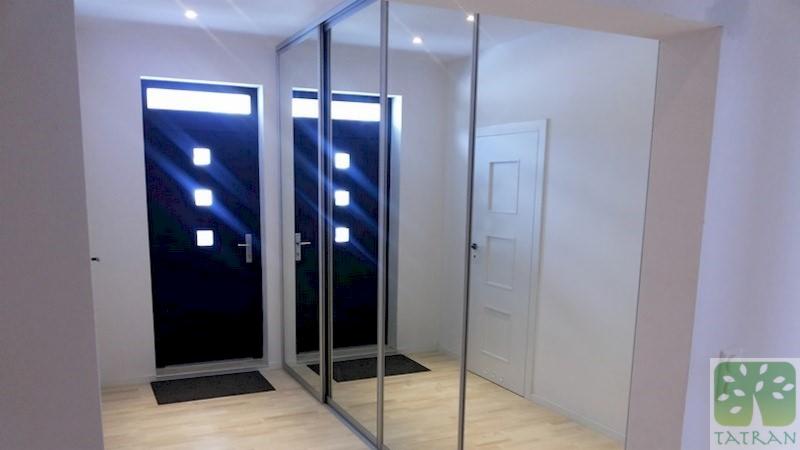 Wnętrze przedpokoju z przesuwną szafą lustrzaną, odbijającą czarne drzwi wejściowe z oświetleniem i białe drzwi wewnętrzne, na jasnej drewnianej podłodze, z logo firmy w prawym dolnym rogu.