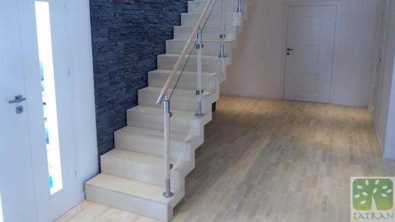 Jasne drewniane schody z balustradą ze szkła i stali nierdzewnej, ściana z dekoracyjnej szarej cegły w tle, widoczny fragment parkietu i dwoje białych drzwi.