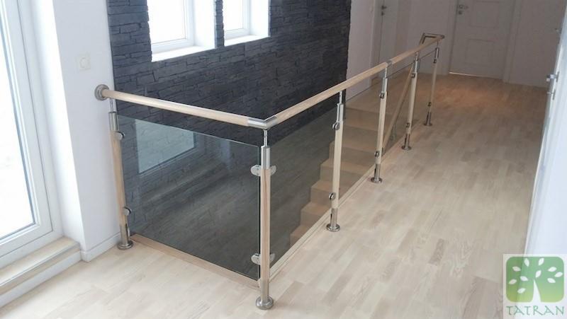 Szklana balustrada schodowa z drewnianymi elementami i metalowymi mocowaniami, widok z góry na jasną podłogę i fragment ściany z dekoracyjną okładziną kamienną.