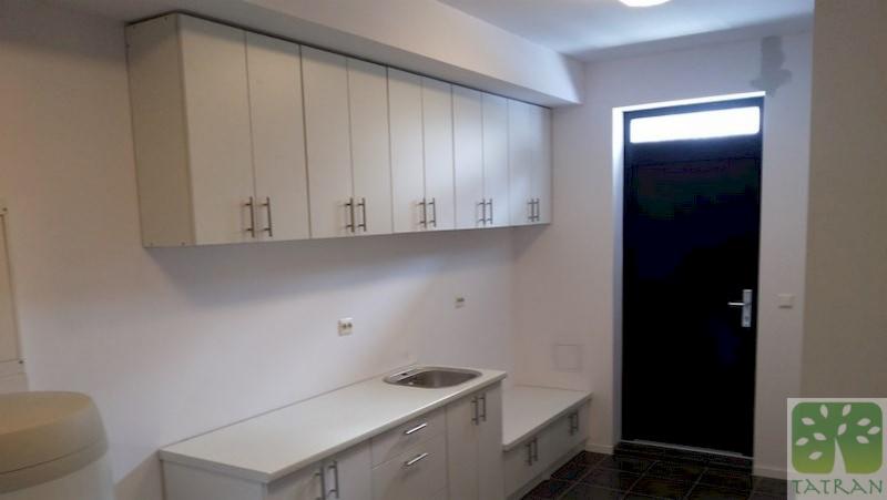 Wnętrze pomieszczenia gospodarczego z białymi szafkami wiszącymi i stojącymi z metalowymi uchwytami, zlewem i czarnymi drzwiami wejściowymi.