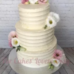 Trzypiętrowy tort śmietankowy z poziomymi prążkami, udekorowany białymi i różowymi eustomami, umieszczony na srebrnej paterze na tle białej ceglanej ściany i drewnianej podłogi.