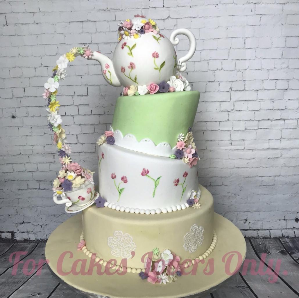 Wielopoziomowy tort zdobiony motywem filiżanki i dzbanka, połączonych kwiatową girlandą, ustawiony na paterze na tle ściany z białej cegły.