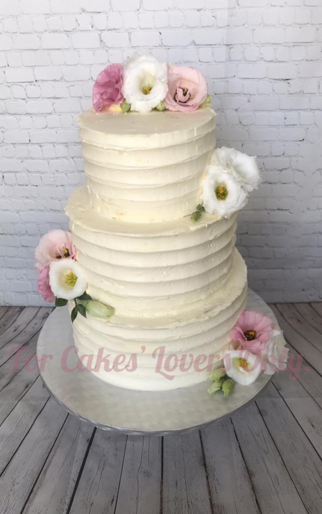 Trzypiętrowy tort śmietankowy z poziomymi prążkami, udekorowany białymi i różowymi eustomami, umieszczony na srebrnej paterze na tle białej ceglanej ściany i drewnianej podłogi.