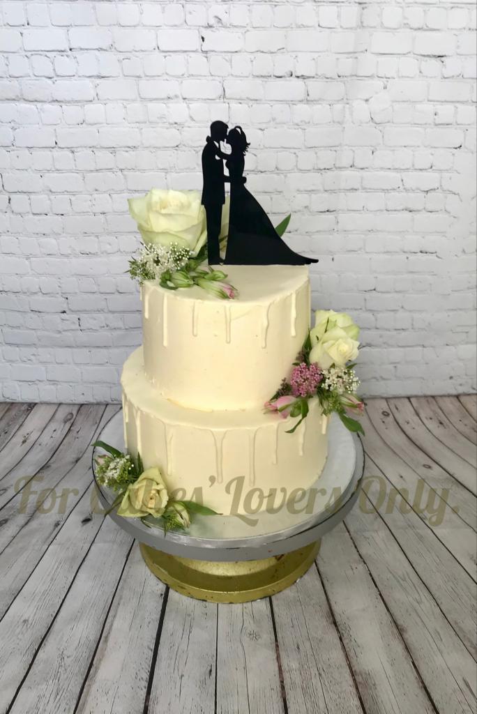 Dwupoziomowy tort weselny z białym kremem, dekorowany kwiatami i topperem z sylwetką pary, na drewnianym stole i tle białej cegły.