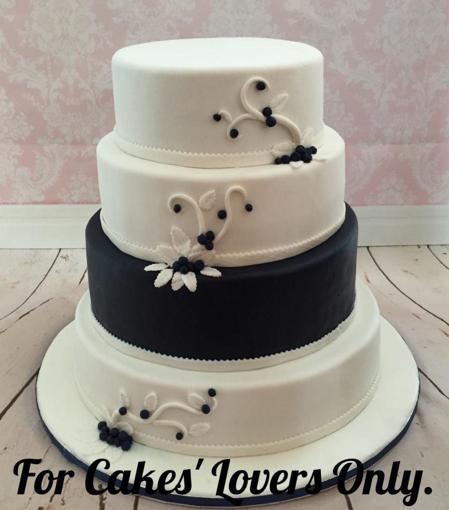 Trzypiętrowy tort weselny w kolorach bieli i granatu, zdobiony kremowymi ornamentami z motywem liści i kulek, umieszczony na drewnianym blacie, z napisem 'For Cakes' Lovers Only'.