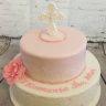 Dwupiętrowy tort w odcieniach różu i bieli, udekorowany kwiatem z lukru i krzyżem na szczycie, z napisem okolicznościowym na dolnym piętrze.