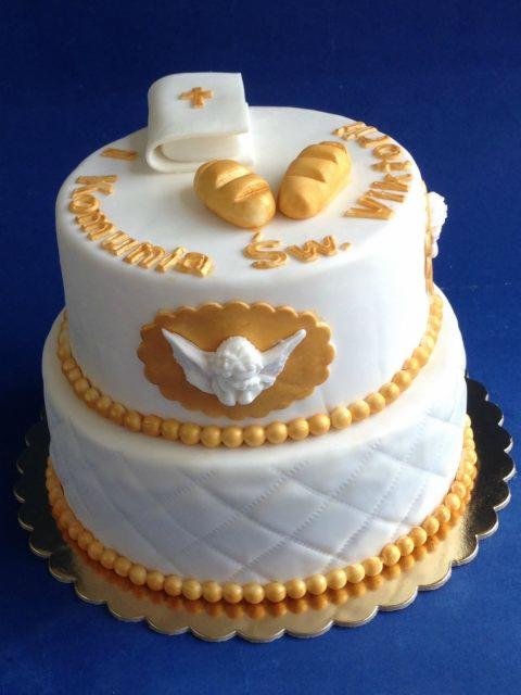 Dwupiętrowy tort komunijny z białym lukrem, złotymi zdobieniami w postaci koralików, aniołka i napisu oraz dekoracjami z lukru imitującymi książeczkę i chlebki.