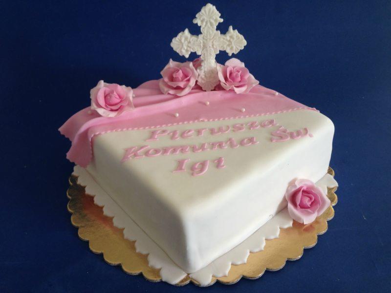 Kwadratowy tort na Pierwszą Komunię Świętą z białym lukrem, różowym napisem 'Pierwsza Komunia Św. Igi', dekoracjami z różowych róż i białym krzyżem na wierzchu, na złotej podkładce.