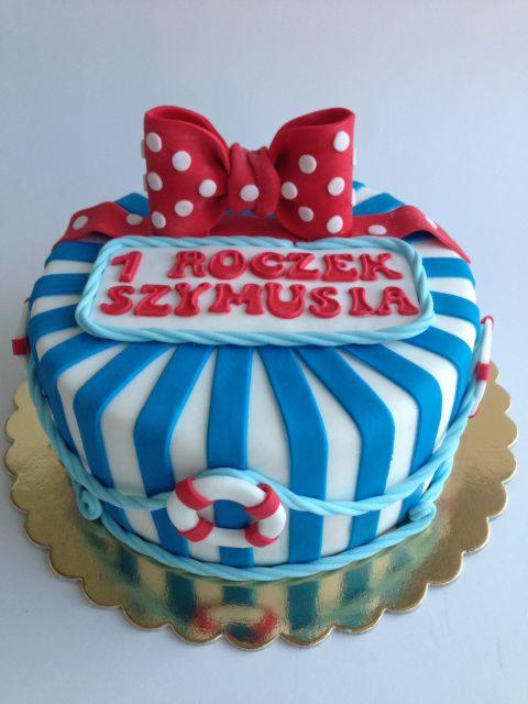 Tort na pierwsze urodziny dziecka w stylu marynistycznym: biało-niebieskie paski, czerwona kokarda w białe kropki i napis '1 Roczek Szymusia' na białej tabliczce.
