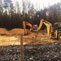 Dwa żółte i pomarańczowe koparki CAT pracują na placu budowy z widocznymi pryzmami ziemi i kamieniami w tle lasu.