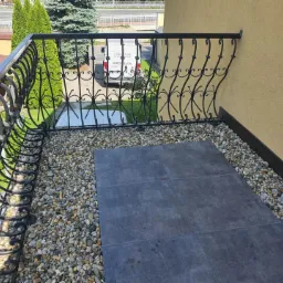 Wykończony balkon z ozdobną, czarną, kutą balustradą, szarymi płytkami podłogowymi i drobnymi kamieniami dekoracyjnymi, widok z góry, jasne światło dzienne.