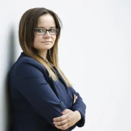 Portret kobiety w okularach i granatowym żakiecie, opierającej się o białą ścianę z założonymi rękami.