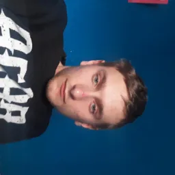 Portret młodego mężczyzny na tle jednolitej, niebieskiej ściany w Brodnicy, ubrany w czarną bluzę z białym nadrukiem.