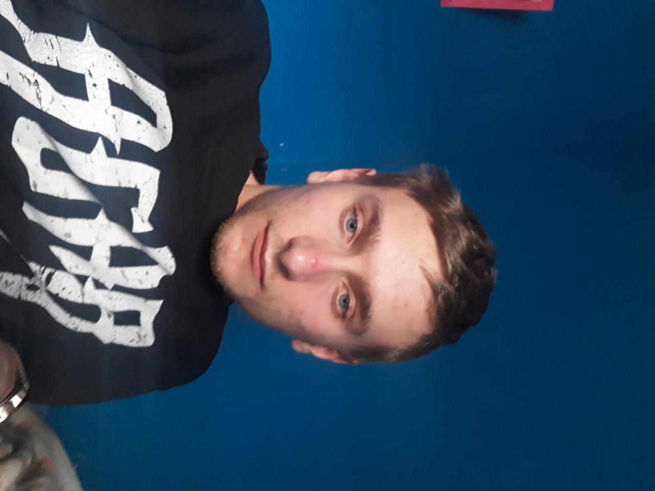 Portret młodego mężczyzny na tle jednolitej, niebieskiej ściany w Brodnicy, ubrany w czarną bluzę z białym nadrukiem.