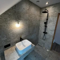 Wykończona łazienka na poddaszu z szarymi płytkami imitującymi beton, białą, prostokątną umywalką nablatową z czarną armaturą i czarnym zestawem prysznicowym.