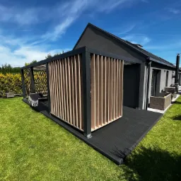 Nowoczesny taras z deski kompozytowej w kolorze antracytowym, z ażurową zabudową z jasnego drewna, przylegający do szarego domu z ciemnym dachem, otoczony zielonym trawnikiem i tujami.