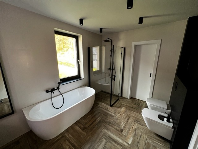 Nowoczesna łazienka z wolnostojącą wanną, kabiną prysznicową typu walk-in z czarnymi ramami, toaletą i bidetem w stylu minimalistycznym, na podłodze parkiet ułożony w jodełkę.