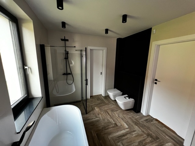 Nowoczesna łazienka z wanną, kabiną prysznicową typu walk-in z czarnymi ramami, toaletą i bidetem na drewnopodobnej podłodze w jodełkę, oświetlona czarnymi reflektorami sufitowymi.