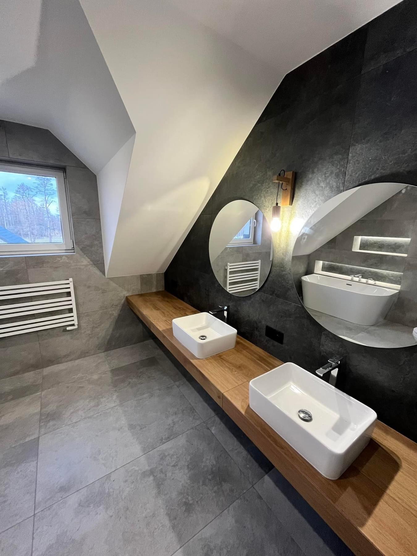 Nowoczesna łazienka na poddaszu z dwoma białymi umywalkami nablatowymi na drewnianym blacie, okrągłe lustra odbijające wannę, szare płytki na ścianach i podłodze, oraz grzejnik drabinkowy.