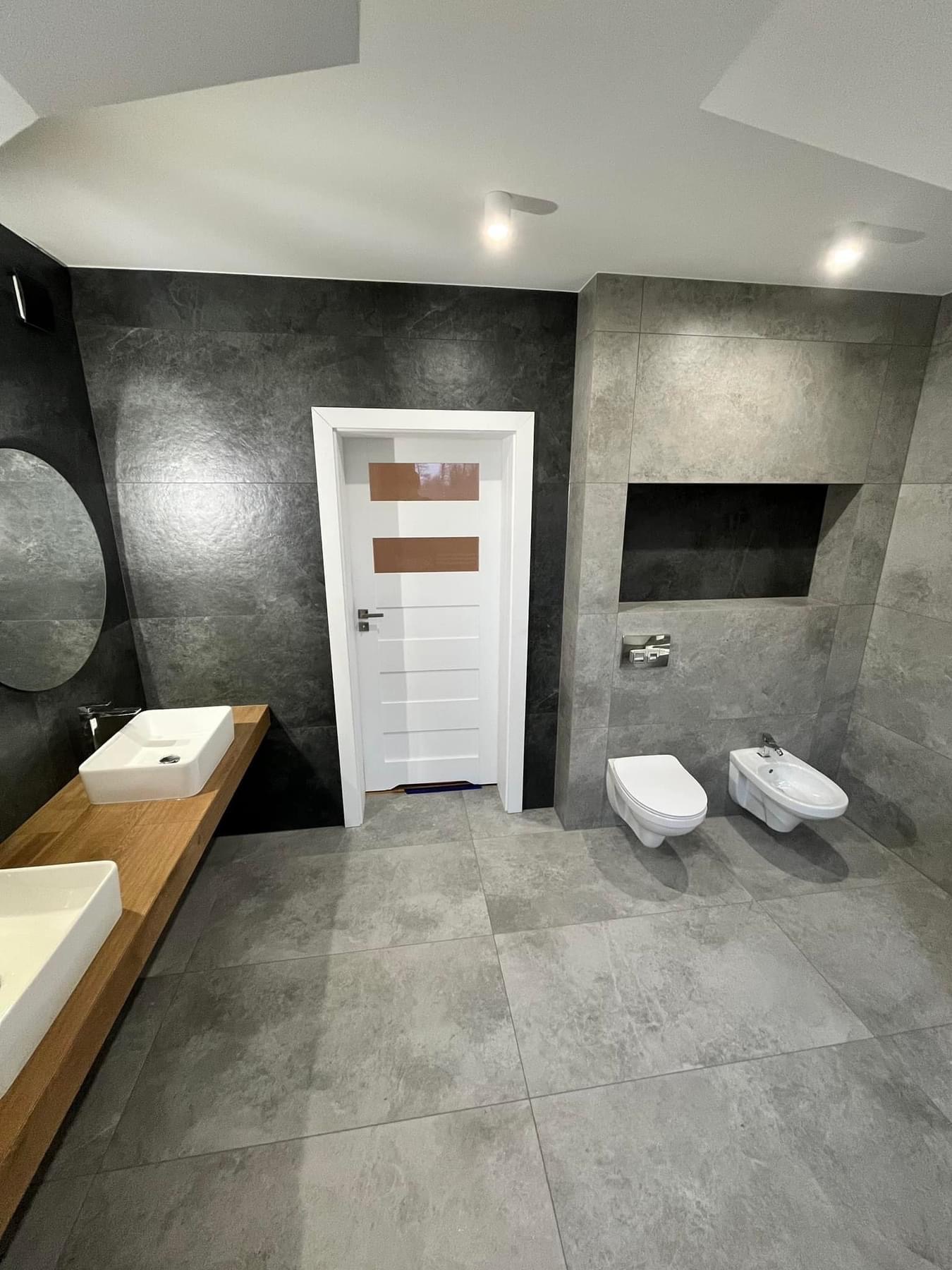 Nowoczesna łazienka z dwoma umywalkami nablatowymi na drewnianym blacie, toaletą i bidetem podwieszonymi na szarej, płytkowej ścianie, białe drzwi z brązowymi wstawkami, oświetlenie punktowe.