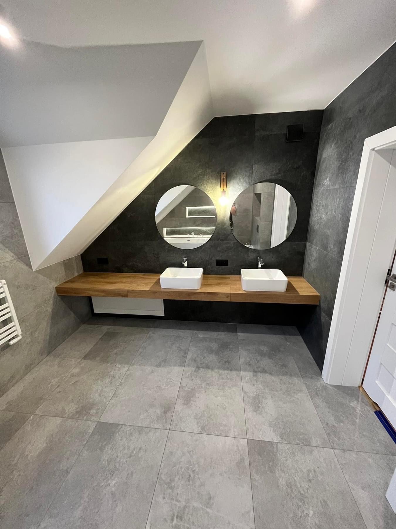 Nowoczesna łazienka na poddaszu z dwoma kwadratowymi umywalkami na drewnianym blacie, okrągłe lustra z podświetleniem LED i szare płytki na ścianach i podłodze.
