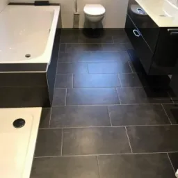 Nowoczesna łazienka z ciemnoszarą, prostokątną glazurą podłogową ułożoną poziomo, białą wanną z ciemnym obramowaniem oraz białą umywalką z szafką w kolorze antracytowym.