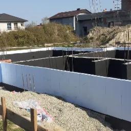 Budowa fundamentów: Szalunek z białych płyt styropianowych, izolacja przeciwwilgociowa w kolorze czarnym, wystające pręty zbrojeniowe, widoczny rów pod fundamenty oraz nasyp ziemi w tle.