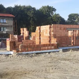 Wznoszenie ścian domu z czerwonej ceramiki Porotherm na warstwie hydroizolacji, widoczne zbrojenie pionowe i palety z pustakami, w tle budynek mieszkalny.