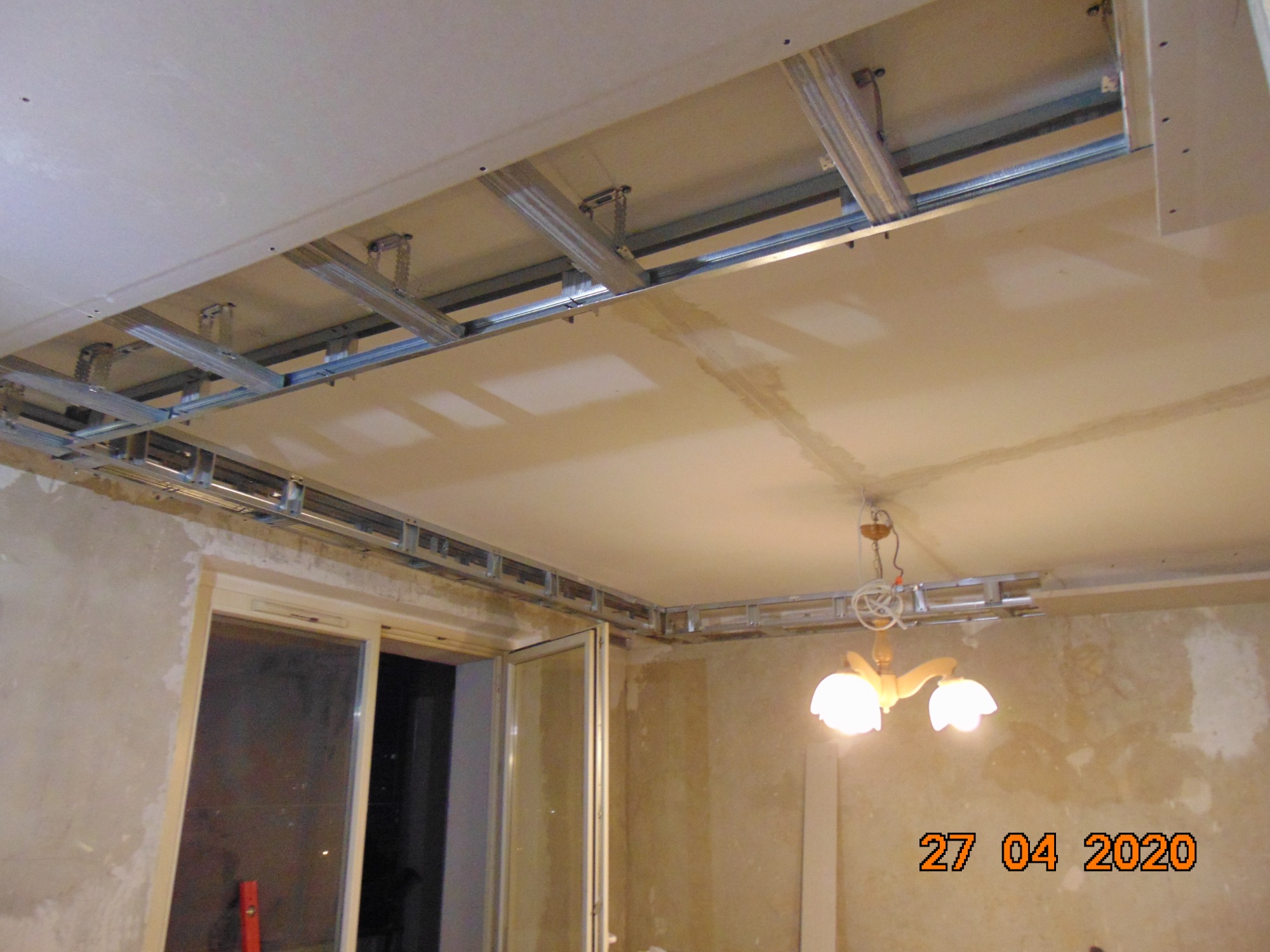 Montaż metalowego stelażu sufitu podwieszanego z płyt gipsowo-kartonowych, widoczny fragment ściany z pozostałościami starej tapety i okno balkonowe.