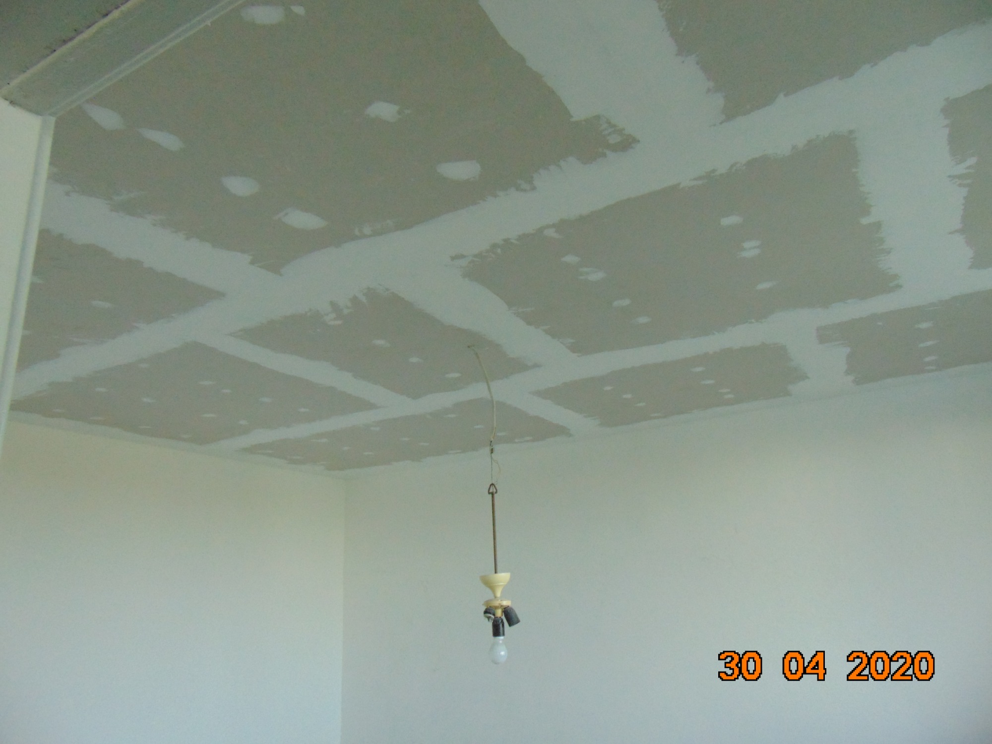 Sufit podwieszany z płyt gipsowo-kartonowych w trakcie szpachlowania łączeń, widoczne niedoskonałości i ślady po wkrętach, surowa żarówka na kablu zwisająca z sufitu, ściany w kolorze złamanej...