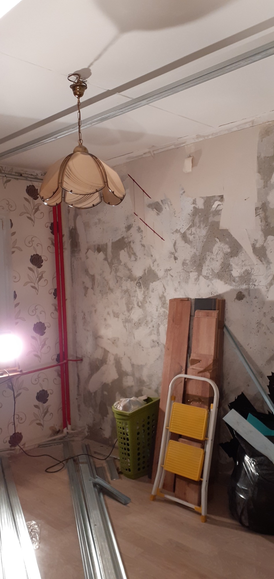 Pokój w trakcie remontu: odsłonięta ściana z resztkami tynku, przygotowane profile metalowe, drabina i lampa wisząca.
