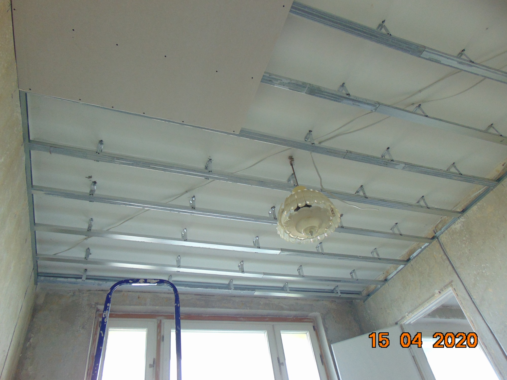 Montaż sufitu podwieszanego z płyt kartonowo-gipsowych na metalowej konstrukcji szkieletowej, widoczny fragment płyty, nieotynkowane ściany, drabina i wisząca lampa.