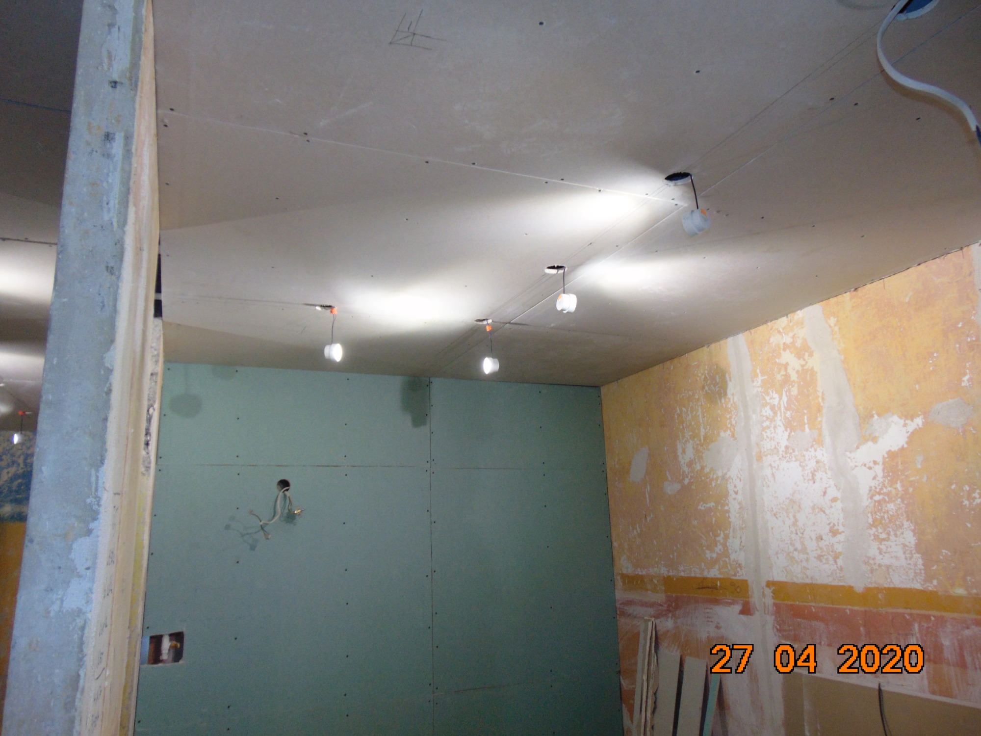 Wnętrze w trakcie remontu: ściany częściowo pokryte płytami gipsowo-kartonowymi, widoczne kable elektryczne, zamontowane tymczasowe oświetlenie, ściany w tle z resztkami starej farby.