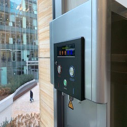 Dozownik wody pitnej z wyświetlaczem, zamontowany w biurze w Piasecznie, z widokiem na nowoczesny budynek przez okno.