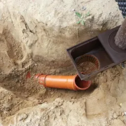 Wykonany wykop z ułożoną pomarańczową rurą drenarską i brązową studzienką drenażową z filtrem, widoczne ślady po wykopie w piasku.