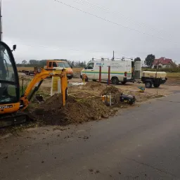 Żółty minikoparka JCB 8014 CTS w trakcie prac ziemnych, z widocznymi przewodami i sprzętem do przewiertów sterowanych, na tle pojazdów serwisowych i krajobrazu wiejskiego.