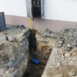 Odkopany rów z ułożoną kostką brukową, widoczne rury w ziemi i skrzynka gazowa na ścianie budynku, zabezpieczona folią.