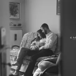 Czarno-białe zdjęcie przedstawia parę siedzącą na kanapie w pokoju, trzymającą noworodka. Kobieta w jasnej bluzce i dżinsach, mężczyzna w koszuli w drobne wzory i ciemnych spodniach. W tle widoczne...